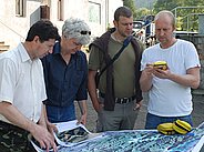 Vier Personen stehen um einen großen, ausgebreiteten Plan. Eine Person hält ein gelbes Gerät in der Hand, während die anderen auf den Plan schauen.