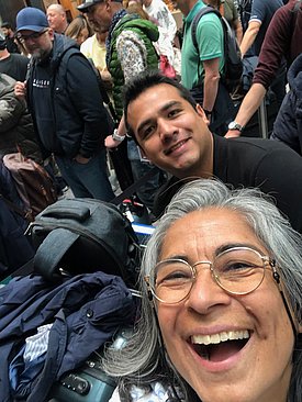 A smiling woman with gray hair and glasses takes a selfie with a man in a black shirt, both looking happy. In the background, a crowd of people is gathered, suggesting a lively event or gathering. The focus is on their cheerful expressions.