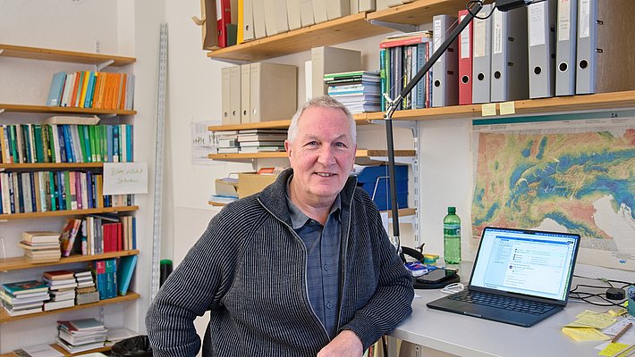 Un homme souriant est assis à un bureau, avec un ordinateur portable ouvert devant lui. En arrière-plan, des étagères remplies de livres et de dossiers sont visibles. La pièce semble être un bureau de travail.