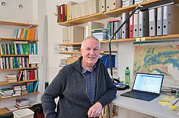 A smiling man sits next to a computer on a desk, with shelves of books and files in the background. He wears a dark sweater and has gray hair, looking comfortably at the camera in an office-like environment.