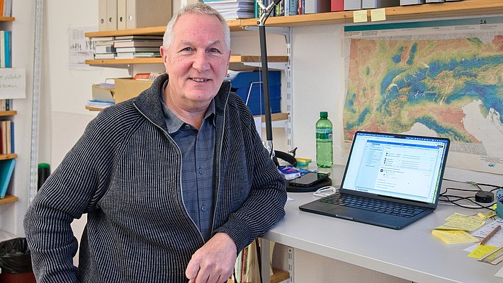 Ein Mann sitztan einem Tisch mit einem Laptop. Im Hintergrund sind Regale mit Ordnern und Büchern zu sehen. Der Raum wirkt akademisch, und auf dem Tisch liegt eine Karte. Der Mann trägt einen grauen Pullover und lächelt.