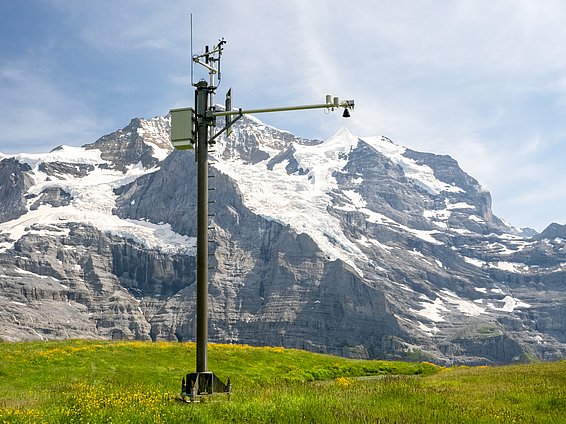 Messstation mit Ultraschallsensor auf grünem Feld vor schneebedecktem Bergmassiv unter blauem Himmel