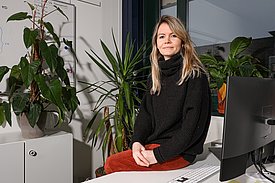 A woman with long blonde hair sits on a desk in an office surrounded by various indoor plants. She wears a black turtleneck sweater and reddish-orange pants. A computer is visible next to her, along with a white board in the background.