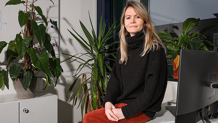 Eine lächelnde Frau mit langen, blonden Haaren sitzt an einem Tisch in einem Raum, der mit einer Vielzahl von Pflanzen dekoriert ist. Sie trägt einen schwarzen Pulli und eine rötliche Hose. Im Hintergrund ist ein Computer und eine Wandtafel sichtbar.