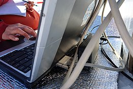 Hand tippt auf Tastatur eines geöffneten Laptops, der auf einer reflektierenden Unterlage steht, mit mehreren Kabeln angeschlossen.