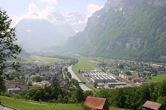Blick auf den Flughafen Mollis mit Start- und Landebahn, umgeben von grünen Wiesen, Gebäuden und hohen Bergen im Hintergrund