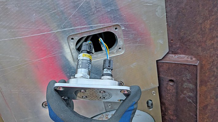 Eine Hand in einem schwarzen Handschuh hält ein metallisches Bauteil mit zwei Sensoren, die Kabel haben. Im Hintergrund ist eine verkratzte Metalloberfläche mit Löchern und Anzeichen von Rost zu sehen.