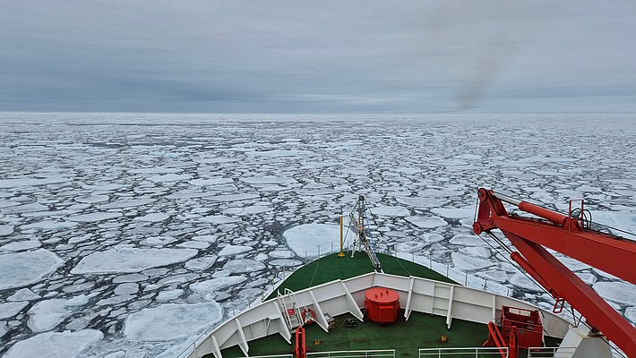 Vorderer Teil eines Schiffes mit grünem Deck und roter Ausrüstung, das durch zerklüftetes Meereis fährt.