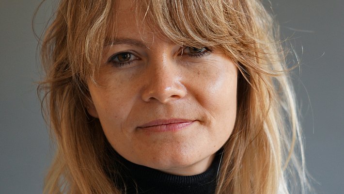 Portrait d'une femme avec des cheveux blonds, vêtue d'un pull noir. Elle regarde l'objectif avec une expression neutre sur fond gris.