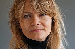 A woman with long, light brown hair and a relaxed expression is looking directly at the camera. She is wearing a black turtleneck sweater. The background is a soft gray color.