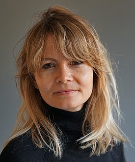 A woman with long, light brown hair and a relaxed expression is looking directly at the camera. She is wearing a black turtleneck sweater. The background is a soft gray color.