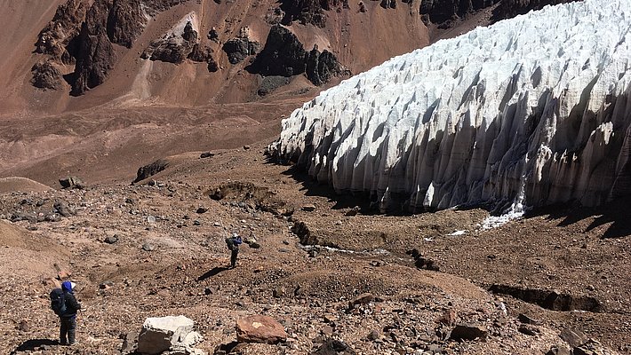 Two hikers stand on rocky terrain, looking towards a large, glacial formation that contrasts with the surrounding arid landscape. The sharp spikes of snow and ice are typical of dry mountain regions. Meltwater streams pour from the glacier. The backdrop features rugged mountains.