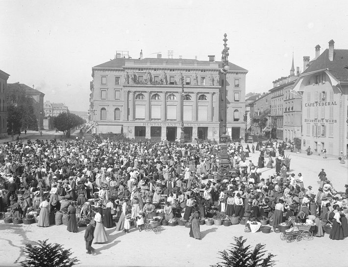 A large crowd gathers in a market square in front of the Federal Palace in Bern. The photo shows the market in 1902.