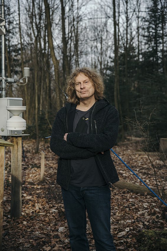 A man with curly hair stands with his arms crossed in front of a forest and a weather station.