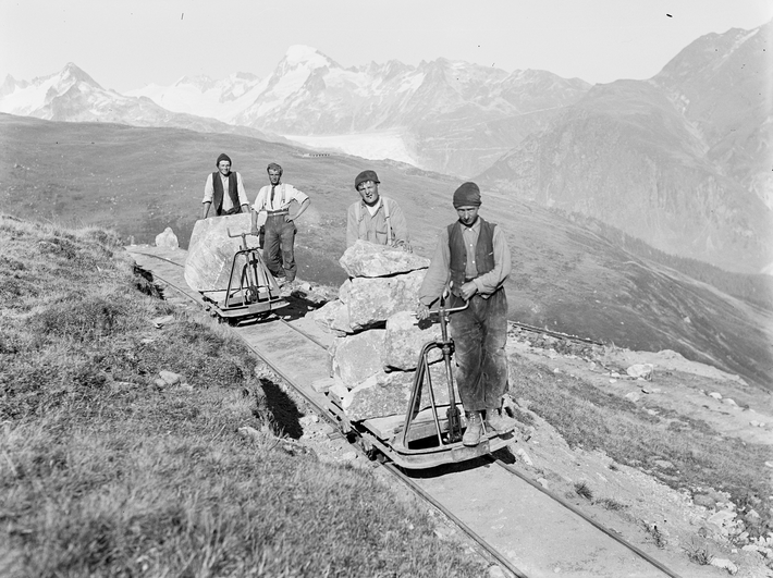 Four workers transport large stones on rail cars through a mountainous landscape. Snow-capped mountains can be seen in the background.