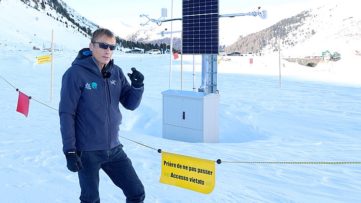 Person mit Sonnenbrille und Winterjacke steht neben einer Messstation mit Solarpanel in verschneiter Berglandschaft
