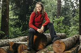 Die neue Einheitsleiterin Janine Schweier sitzt auf einem Holzstapel im Wald. Sie trägt eine rote Jacke und schwarze Hose. Im Hintergrund sind hohe Bäume und grünes Unterholz sichtbar. Die Stimmung wirkt naturalistisch und entspannt.