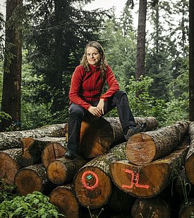 Die neue Einheitsleiterin Janine Schweier sitzt auf einem Holzstapel im Wald. Sie trägt eine rote Jacke und schwarze Hose. Im Hintergrund sind hohe Bäume und grünes Unterholz sichtbar. Die Stimmung wirkt naturalistisch und entspannt.