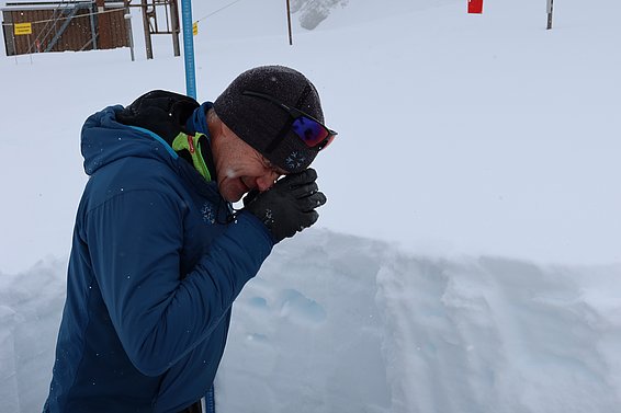 Person in blauer Winterjacke und schwarzer Mütze benutzt Lupe zur Schneekristallbestimmung im Schnee