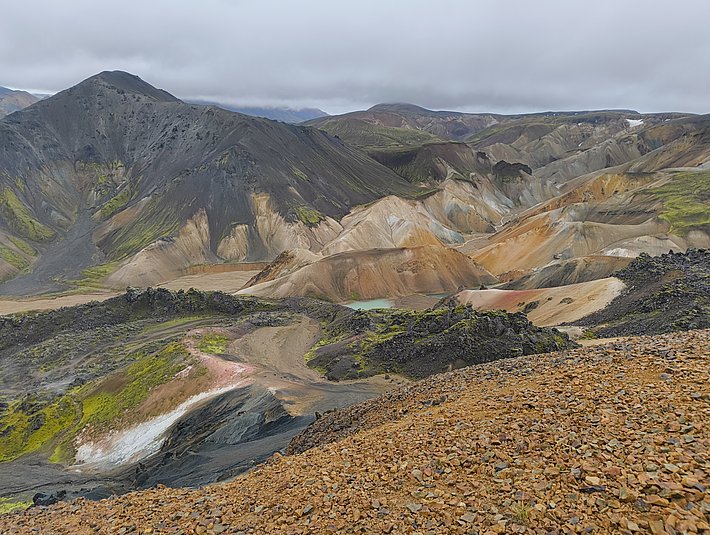 Eine weite Landschaft mit bunten Bergformationen in verschiedenen Erdetönen, darunter Grau, Braun und Ocker. Über die Szenerie ziehen graue Wolken, und im Vordergrund sind steinige Flächen und einige grüne Pflanzen zu sehen.