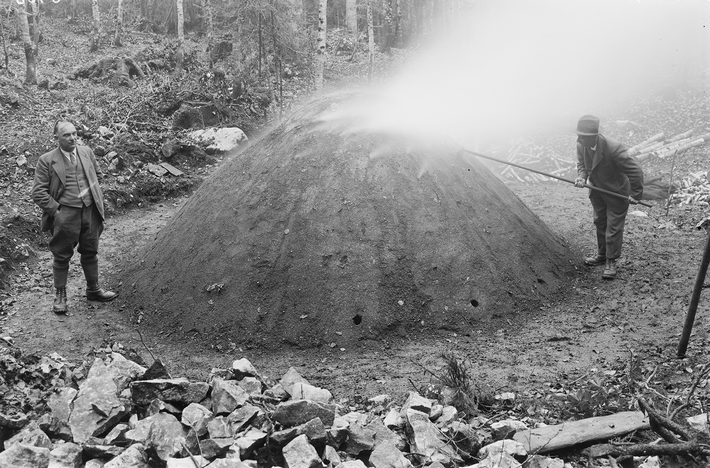 Two men are standing in a wooded area next to a large hill. One of the men is poking around with a long stick in the hill, from which smoke is rising.