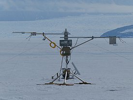 Ein wissenschaftliches Messgerät steht auf einer verschneiten Fläche, umgeben von Eis. Es verfügt über mehrere Sensoren und Antennen, die zur Datenerfassung in einer polarähnlichen Umgebung verwendet werden. Im Hintergrund sind schneebedeckte Berge und ein eisiger Himmel zu sehen.