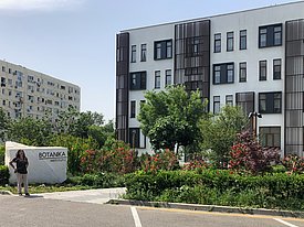 Carolina stands next to a stone sign reading "Botanika Institute," in front of a modern building surrounded by greenery and colorful plants.