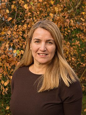 A woman with long, blonde hair smiles directly at the camera, wearing a dark brown top. She is surrounded by autumn foliage, featuring warm tones of orange, yellow, and brown, creating a natural and inviting backdrop.