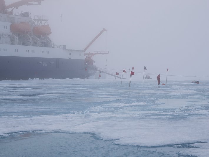 Ein Forschungsschiff mit orangefarbenen Kränen steht im Nebel. Auf dem Eis sind mehrere Flaggen und eine Person in roter Kleidung zu sehen.