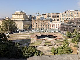 Blick auf eine kreisförmige, teilweise unterirdische Struktur mit Wegen und Grünflächen umgeben von mehrstöckigen Gebäuden in einer Stadt.