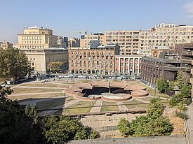 A view of a circular, partly underground structure with paths and green spaces, surrounded by multi-storey buildings in a city.