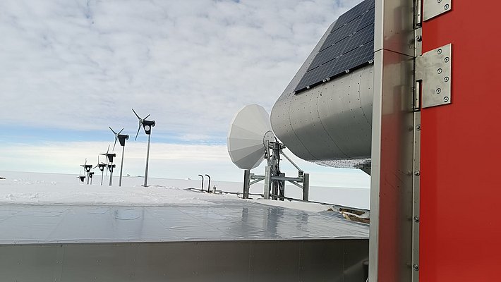 Reihe kleiner Windturbinen auf verschneitem Gelände mit einem großen Solarpanel und einer Satellitenschüssel im Vordergrund