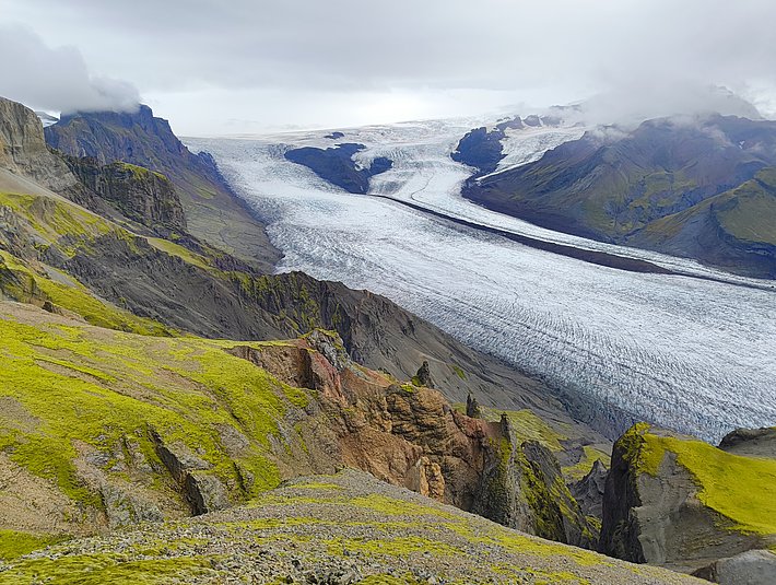 Eine weitläufige Landschaft mit dramatischen Felsformationen umgeben von sattem Grün. In der Mitte fliesst ein grosser Gletscher, der sich bis in die Ferne erstreckt. Über dem Gletscher hängen graue Wolken, die eine mystische Atmosphäre schaffen.