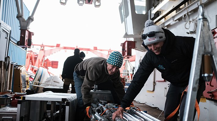 Zwei Personen in Arbeitskleidung und Mützen greifen auf einem Schiff nach Ausrüstungsteilen in einem offenen Laderaum mit Containern und Kran im Hintergrund.