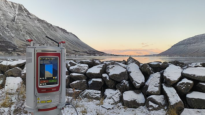 Ein elektronisches Messgerät steht auf einem Stativ vor einer schneebedeckten Steinmauer. Im Hintergrund erstrecken sich schneebedeckte Berge und ein ruhiger See, während der Himmel in sanften Farben der Dämmerung erleuchtet ist.