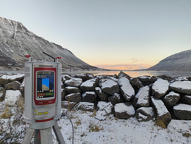 Ein elektronisches Messgerät steht auf einem Stativ vor einer schneebedeckten Steinmauer. Im Hintergrund erstrecken sich schneebedeckte Berge und ein ruhiger See, während der Himmel in sanften Farben der Dämmerung erleuchtet ist.
