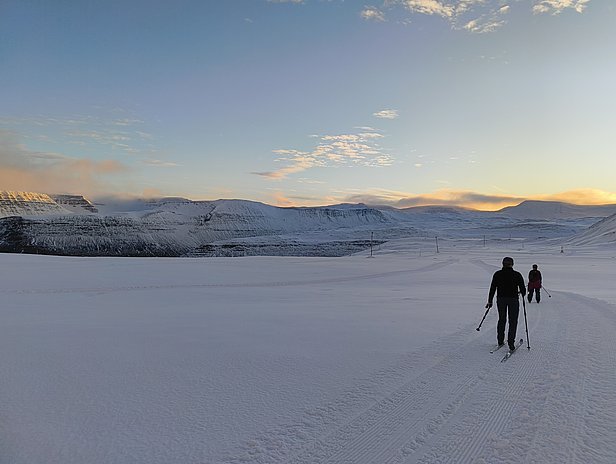 Zwei Skifahrer bewegen sich durch eine schneebedeckte Landschaft. Im Hintergrund sind majestätische Berge und ein heller, klarer Himmel zu sehen. Die Szene vermittelt eine ruhige, winterliche Atmosphäre.