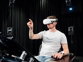 Mann mit weißem VR-Headset und Controller in einem dunklen Raum mit Lautsprechern, hebt eine Hand zum Interagieren.