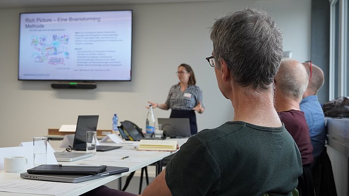 Eine Präsentation in einem Seminarraum wird gezeigt. Im Vordergrund sitzen Teilnehmer, die einem Vortrag folgen. Eine Person steht vor einem Bildschirm und erklärt Visualisierungen und Text, die auf dem bildschirm zu sehen sind.