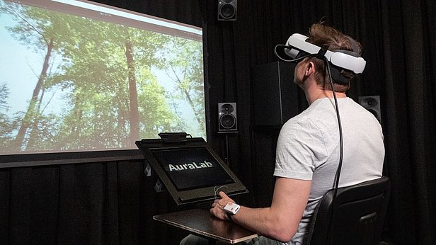 Mann mit VR-Brille sitzt vor Bildschirm mit Waldansicht, vor ihm ein Tablet mit Schrift 'AuraLab' in dunklem Raum.