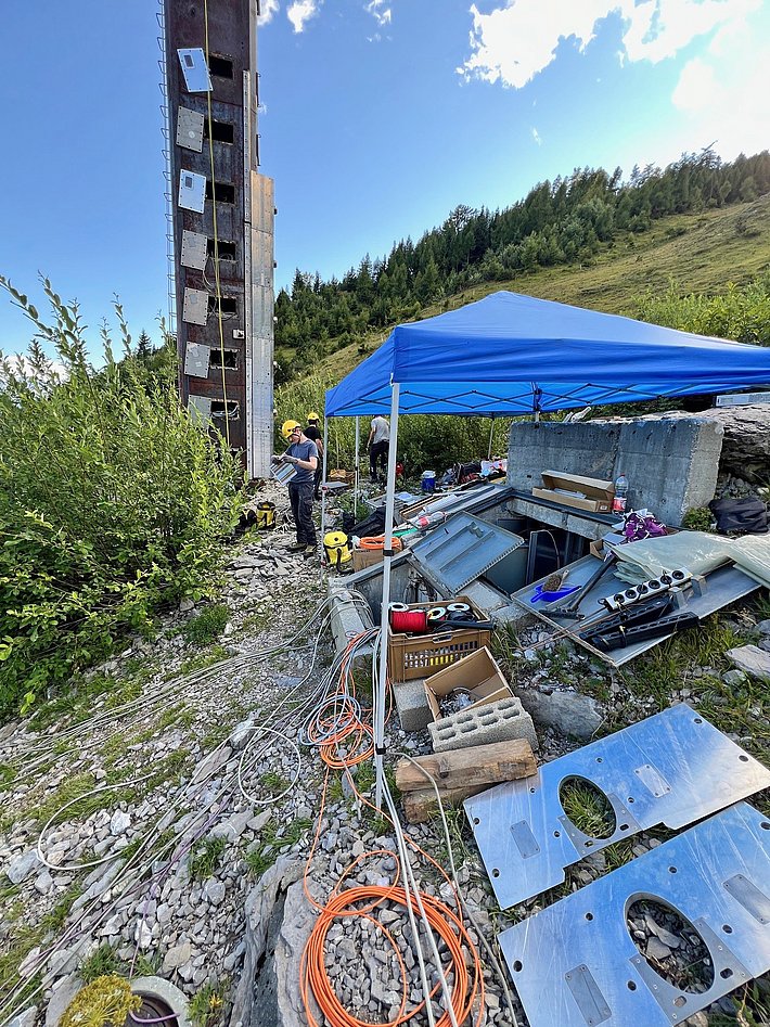 Eine kleine Baustelle im Freien mit einem hohen Mast im Hintergrund. Ein blauer Pavillon steht auf dem Boden, umgeben von Werkzeugen, Materialien und Kabeln. Im Vordergrund sind verschiedene Metallteile und Kisten zu sehen, während Arbeiter mit Installationen beschäftigt sind.
