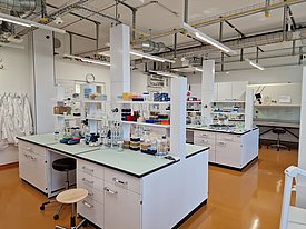 A modern laboratory with multiple workbenches, glassware, and equipment arranged neatly. Lab coats hang on hooks, and shelves display various containers and tools. The floor is orange, and overhead pipes and lighting fixtures are visible. The environment appears clean and organized.