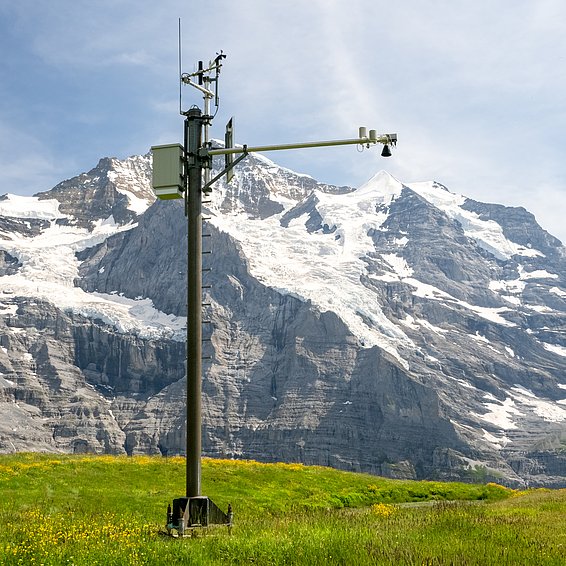 Messmast mit Sensoren auf einer grünen Wiese vor schneebedeckten Bergen unter blauem Himmel.