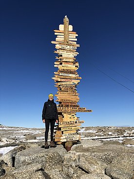 Eine Frau steht neben einem Schilderbaum und lächelt in die Kamera. Sie ist warm angezogen und hinter ihr sind Schneereste zu sehen. Der Himmel ist strahlend blau.