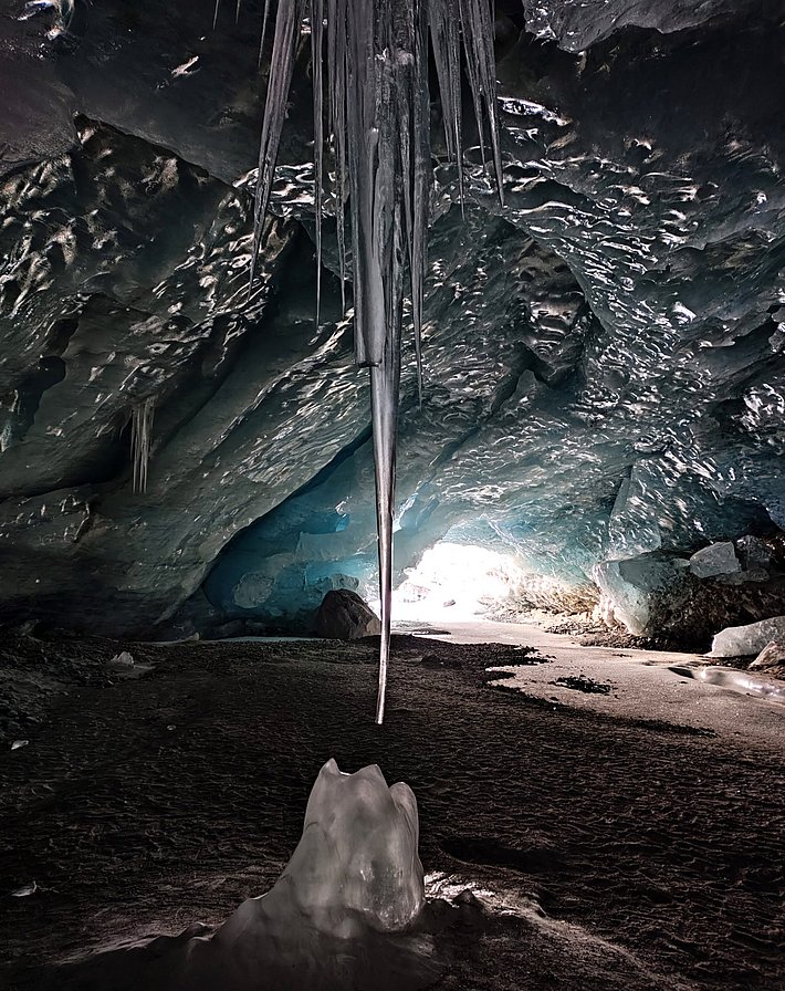 Long, pointed icicles hang from the ceiling of a dark ice cave with an uneven floor and a bright entrance in the background.