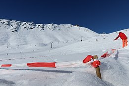 Schneebedeckte Versuchsfläche mit rot-weißem Absperrband im Vordergrund und Person in roter Jacke am Rand, im Hintergrund schneebedeckte Berge unter blauem Himmel