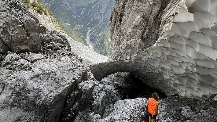 A person in an orange shirt stands on rocky terrain, looking up at a large, textured rock formation resembling a natural arch. Majestic mountains rise in the background under a cloudy sky, highlighting a remote and rugged landscape.