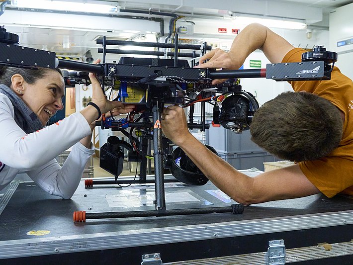 Zwei Personen montieren eine große Drohne mit schwarzem Rahmen und mehreren Rotorarmen auf einem Tisch in einer Werkstatt.