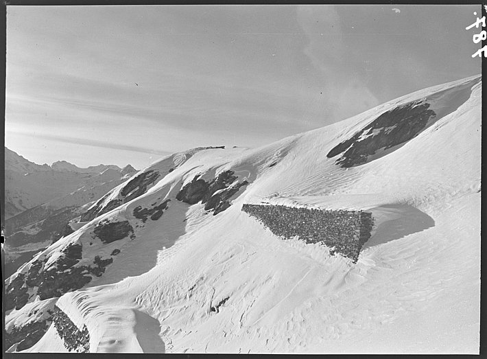 Schneebedeckter Berghang mit mehreren steinernen Stützmauern und felsigen Stellen unter bewölktem Himmel