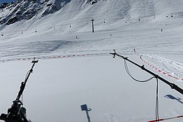 Zwei schwarze Sensorarme mit Kabeln auf einer schneebedeckten Fläche in den Bergen, abgesperrt durch rot-weiße Bänder, im Hintergrund Skifahrer und ein Sessellift